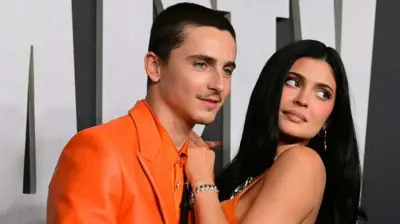 Turuncu takım elbiseli genç bir adam. Yanında uzun siyah saçlı bir kadın. Kameralara bakıyorlar.