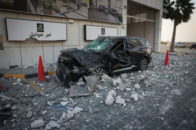 Um carro preto destruído está parado entre dois cones de trânsito vermelhos em meio aos destroços em frente a um hotel em Manama, Bahrein, no domingo, 1º de março de 2026, com outros dois carros atrás de uma palmeira ao fundo.