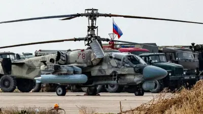Askeri helikopter yanında iki askeri kamyon havaalanında park edilmiş, aralarında bir Rus bayrağı görünüyor. 