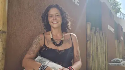 Priscila Albuquerque sorrindo num dia ensolarado, ela tem cabelos cacheados, veste uma regata preta e usa colar e pulseiras de artesanato