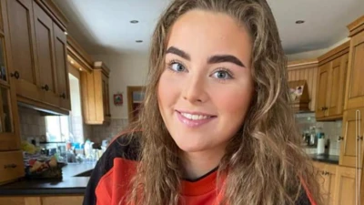 Jeni Larmour smiling in a kitchen. She's wearing a red and black sweatshirt and has light brown curly hair with dark eyebrows. 