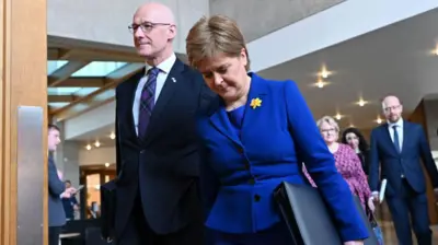 John Swinney and Nicola Sturgeon walking together at the Scottish Parliament. Both are in blue suits and she is carrying a folder There are other people in the background.