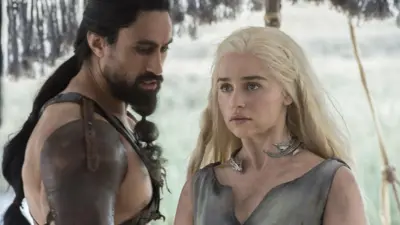 「ゲーム・オブ・スローンズ」の登場人物のジョー・ナウファフとエミリア・クラークが、中世風ファンタジーの革衣装をまとっている。背後には雪が積もっているように見えるが、二人は肌の広い部分を露わにしている。肌は汚れている