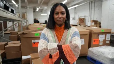 Handbag designer Sherrill Mosee, wearing a bright orange turtleneck, stands with her arms crossed in a warehouse with boxes in the background