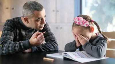 رجل يجلس إلى طاولة ممسكًا قلمًا وينظر إلى طفلة صغيرة تغطي وجهها بيديها، بينما يجلسان أمام كتاب مفتوح، في مشهد يوحي بلحظة توتر أو انفعال أثناء حلّ الواجب المدرسي.