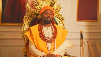 His Majesty, Ogiame Atuwatse III, Olu of Warri sitdown for im royal chair wey be red and get golden design. E wear di traditional red beaded crown wit cream color long sleeve traditional shirt and yellow flowing robe ontop. Red traditional beads dey im neck and wrist, e also carry horsetail for hand and im sword dey for di background.