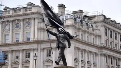 Uma estátua cinza de um homem vestindo um terno, caminhando para frente, carregando uma bandeira que cobre seu rosto 