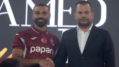 Mohamad Salah ve Trabzonspor Başkanı Ertuğrul Doğan yan yana duruyor ve el sıkışıyorlar. Salah'ın üzerinde Trabzonspor forması var. Doğan ise siyah takım elbise ve beyaz gömlek giyiyor