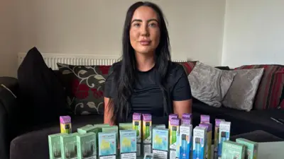 Cara is a 25-year-old woman with long dark hair, she is wearing a black T-shirt and she is sitting on a sofa. On a table in front of her is an array of multi-coloured disposable vape boxes.