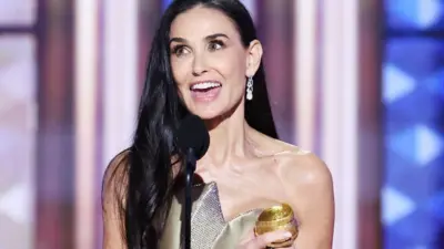 Actress Demi Moore clutches her Golden Globe award to her chest as she makes her thank you speech. She has long dark hair which extends as far as her elbows. and wears a gold dress with bare shoulders. One ear is exposed, from which hangs an ornate earring.