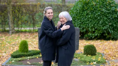 Mezarlıkta gülmesini engellemeye çalışan bir kadın ve yanında gülen bir başka kadın. Anne kız gibi görünüyorlar.