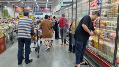 Pessoas no corredor de um supermercado, com gôndolas de vegetais à esquerda e geladeiras de laticínios à esquerda