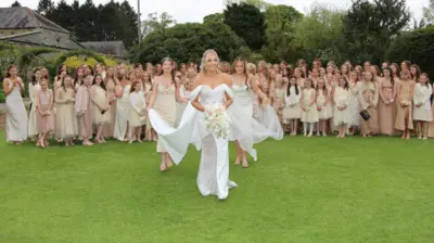 A bride, with blonde hair, a white dress with an off the shoulder detail carrying a bouquet, smiles as two young bridesmaids help her with her train and veil as she walks towards the camera. Behind them is a large group of women, including young girls in the front row, wearing a mixture of champagne, cream, white and tan dresses. The crowd, including the bride and the two bridesmaid, are standing in the middle of a large garden, with tall trees and a leafy tunnel to the left.