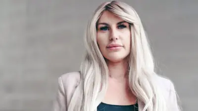 Rebecca posa diante de um muro de pedra clara. Ela tem cabelos loiros longos e ondulados, veste uma blusa preta e uma jaqueta rosa-clara e olha para a câmera com expressão pensativa