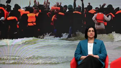 BBCのテレビスタジオに座るマムード内相。背景には、海を小型ボートで渡る人たちが蛍光オレンジの救命胴衣を着け、海辺で波間の中に立つ画像が映し出されている