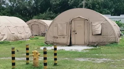 Photo shows three round beige tents on green grass. In the background there is a hut