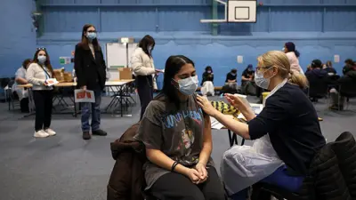 Bir kadın öğrenci kolunu açmış bir kadın hemşire ona aşı yapıyor. Kapalı spor salonu gibi bir alanda küçük masalarda çok sayıda aşı yapılıyor. İki kadın öğrenci ayakta sıra bekliyor.