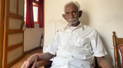 Jageshwar Prasad Avadhiya, egy idős indiai férfi, egy fából készült széken ül. Fehér inget visel, ősz haja és bajusza van.