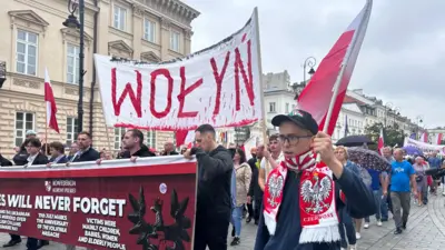 Вулична хода в центрі міста: учасники несуть польські прапори та великий банер із написом "Волинь". На передньому плані юнак із польським прапором і червоно-білим шарфом. Позаду видно натовп, банер англійською мовою про пам’ять жертв, а вдалині інший банер – із перекресленими українським гербом.