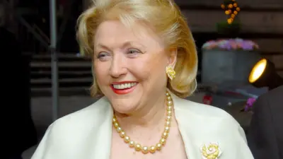 Barbara Taylor Bradford looks to the left of camera while smiling, with blonde hair and a pearl necklace and white suit, pictured in New York in 2000