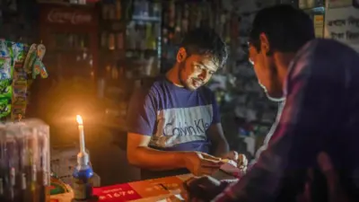 একটি দোকানে মোমবাতি জ্বালিয়ে কাজ করছেন একজন, সামনে আরেকজন দাঁড়িয়ে আছেন