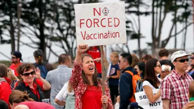 Manifestação contra as vacinas, com uma jovem de vermelho com o punho para o alto, segurando um cartaz dizendo "não à vacinação forçada", em inglês.