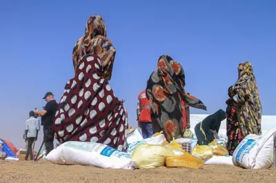 يظهر في الصورة المتلقطة في 25 نوفمبر/تشرين الثاني 2025، نحو خمس نساء لا تظهر وجوههن في الصورة المأخوذة من الخلف، نزحوا من الفاشر.يرتدين زياً سودانياً ملوناً يغطي رؤوسهن وأجسادهن. هناك أكيس من المساعدات على أرض رملية، وخيام وعدد آخر من الأشخاص في الصورة المتلقطة نهاراً. 
