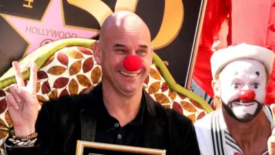 Guy Laliberté attends the Ceremony honouring him with a star on the Hollywood Walk of Fame on 22 November. He is wearing a red clown’s nose and has his hands in a V- sign. To his left is a male clown with a white face paint, red nose, and a black beard and moustache painted into his face. The Hollywood Star is on a plaque behind them and Laliberté is holding a picture frame with the star inside.
