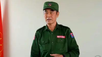 စစ်စိမ်းရောင်ဝတ်စုံကိုဝတ်ဆင်ထားပြီး မတ်တပ်ရပ်အနေအထားနဲ့စကားပြောနေတဲ့ ကိုးကန့်တပ်ခေါင်းဆောင် ဖုန်တာရွှင် ကိုတွေ့ရပါတယ်။
