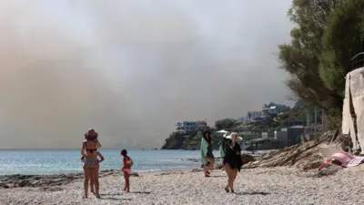 Zadymione niebo podczas pożarów lasów i terenów rolnych w Plomari, na wyspie Lesbos w Grecji. Na pierwszym planie plaża i kilka osób w strojach kąpielowych. 29 lipca 2026 r.