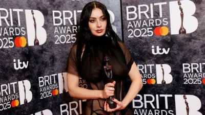 Charli XCX in her sheer dress holding a Brit Award in front of a Brits-branded backdrop