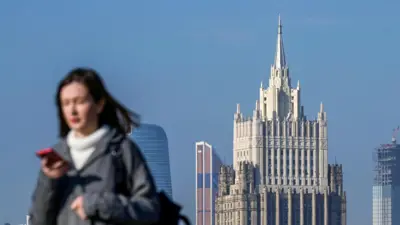 На заднем плане многоэтажное здание со шпилем на фоне голубого неба. На переднем плане в левой части размытая фотография девушки по пояс. Девушка идет справа налево. У нее темные длинные волосы. Она одета в серую куртку и белый свитер. На левом плече у девушки черная сумка. В правой руке - красный смартфон.