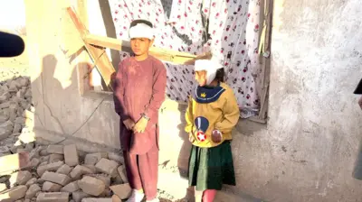 کودکانی که در حملات هوایی پاکستان در پل‌چرخی زخمی شده‌اند سرشان با بانداژ پیچانده شده و در مقابل کلکین یک خانه ویران در مقابل کمره ایستاده‌اند. ۱۳ مارچ ۲۰۲۶ کابل، پل‌چرخی