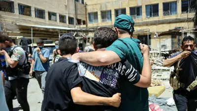Un trabajador sanitario y otros hombres caminan por el patio de un hospital, junto a los cadáveres de las víctimas de los recientes enfrentamientos en la ciudad meridional siria de Sweida, el 17 de julio de 2025.