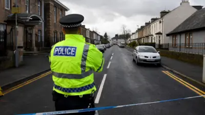 Um policial usando um colete de alta visibilidade atrás de uma fita de isolamento azul e branca que bloqueia uma rua residencial em Glasgow
