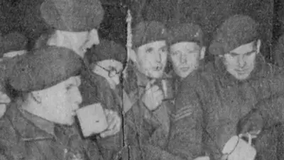 A grainy black-and-white photograph from a newspaper showing paratroopers gathered in a group drinking tea. Each is wearing their beret. It shows a moment just before they got into planes to be dropped over German lines at the River Rhine in Operation Varsity, March 1945.