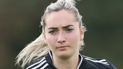 A young woman wearing a black football shirt jogs forward with a determined look on her face. Her long blonde hair is tied back.