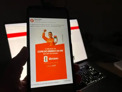 Imagem mostra uma mão segurando um telefone celular num cenário escuro e, na tela, tem um anúncio de uma casa de apostas, a Betano, chamando o usuário para aposta durante a competição
