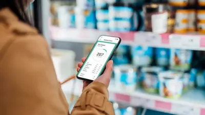 Osoba o niewidocznej twarzy stoi przed półką sklepową z nabiałem. W dłoni trzyma smartfon z aplikacją pokazującą wartość kaloryczną produktu. 