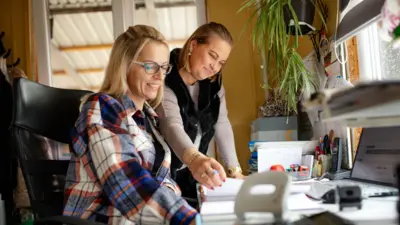 Profesjonalne współpracowniczki biurowe: W harmonijnym środowisku pracy kobieta w średnim wieku, ubrana w koszulę w kratę i okulary, zajmuje się zadaniami finansowymi przy biurku, wspierana przez koleżankę w szarym swetrze i czarnej kamizelce z futerkiem. Zsynchronizowane działania kobiet przyczyniają się do sprawnego funkcjonowania biura i tworzą atmosferę wzajemnej więzi.