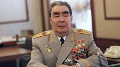 Похилого віку Брежнєв сидить за столом у кабінеті, одягнений у світлий військовий мундир із темною краваткою та численними медалями, орденами та кольоровими планками; на плечах видно оздоблені погони.
На тлі — розмитий інтер’єр із шторами, меблями та телефоном праворуч.