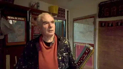 Dan MacNee stands in his home with items stuck on the walls behind him - a map, cut out from a magazine other bits of paper taped to a door and the walls and a bit of wood leant against the wall. He wears an orange sweatshirt over a dark t-shirt and checked shirt, with a paint splattered overall over the top. 