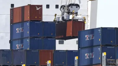 A crane lifts red and blue shipping containers from vessel