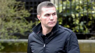 Steven Lyons has cropped brown hair and is wearing a black jacket with the collar turned up. He is looking off to the left with a serious expression on his face