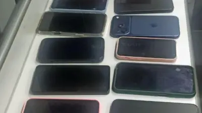 Foto de vários smartphones dispostos sobre uma mesa branca, organizados em fileiras e com as telas apagadas. Há aparelhos de diferentes tamanhos, cores e modelos, incluindo alguns com capinhas. O enquadramento é feito de cima.