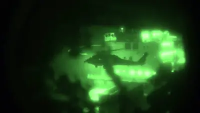 Helicóptero visto con visión nocturna, iluminado en verde, volando cerca de la cubierta de un barco durante la noche.