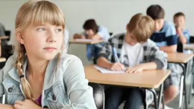 A school classroom scene with a blonde girl gazing to the left and three boys sitting and writing at desks in the rows behind her.