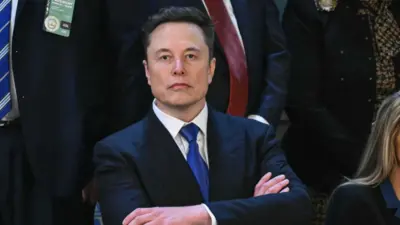 Elon Musk wearing a suit with a blue tie with his arms crossed