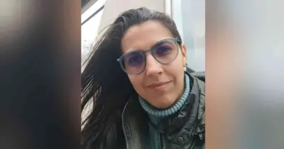 Vitória is looking at the camera up close. She is wearing a blue turtle neck jumper, a khaki or dark-coloured coat, has glasses on, and has brown hair.