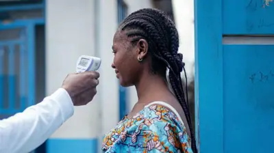 Um médico verifica a temperatura de uma mulher usando um termômetro infravermelho sem contato. Somente o braço do médico, que está vestido de branco, é mostrado. A mulher que está com a temperatura verificada tem o cabelo trançado. Ela está vestindo uma blusa azul brilhante com um padrão e está ao lado de uma porta de madeira azul com um prédio branco e azul ao fundo.